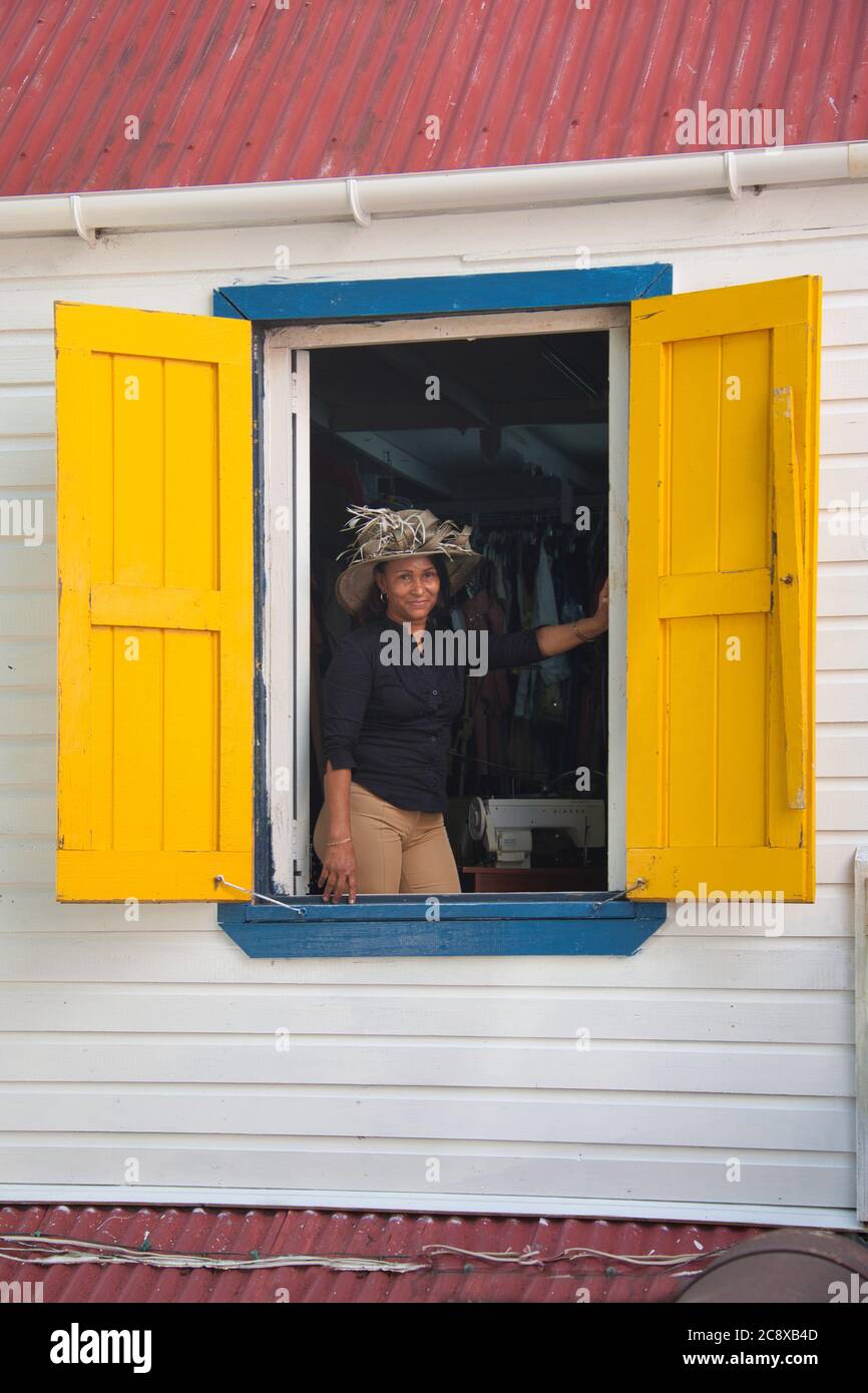 Una signora attraente in un cappello molto fiorito pone per la macchina fotografica in una finestra aperta gialla chiusa a St Johns, isola di Antigua, i Caraibi Foto Stock