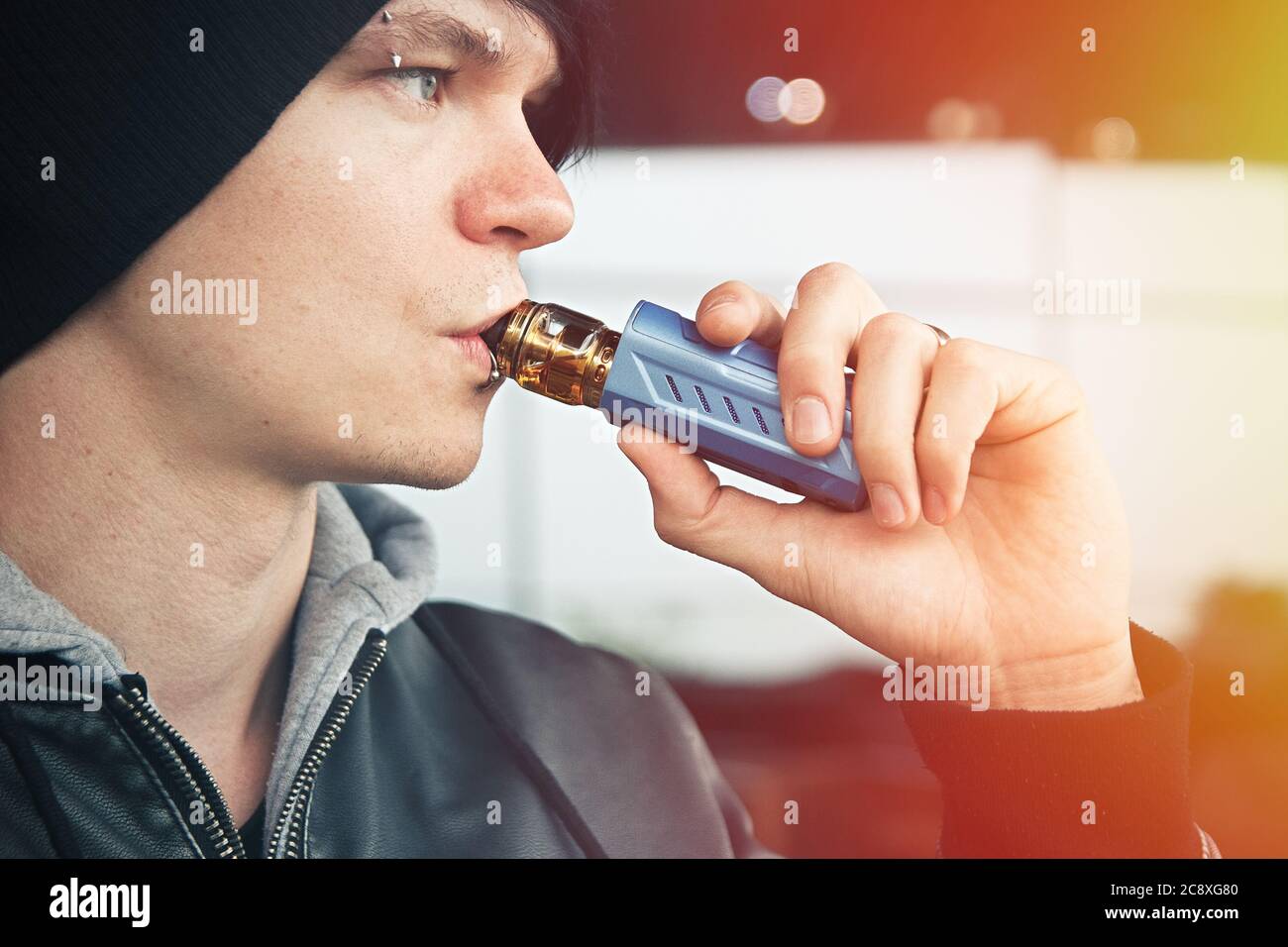 Giovane uomo con cappello e giacca in pelle con cappuccio che fuma un vape Foto Stock