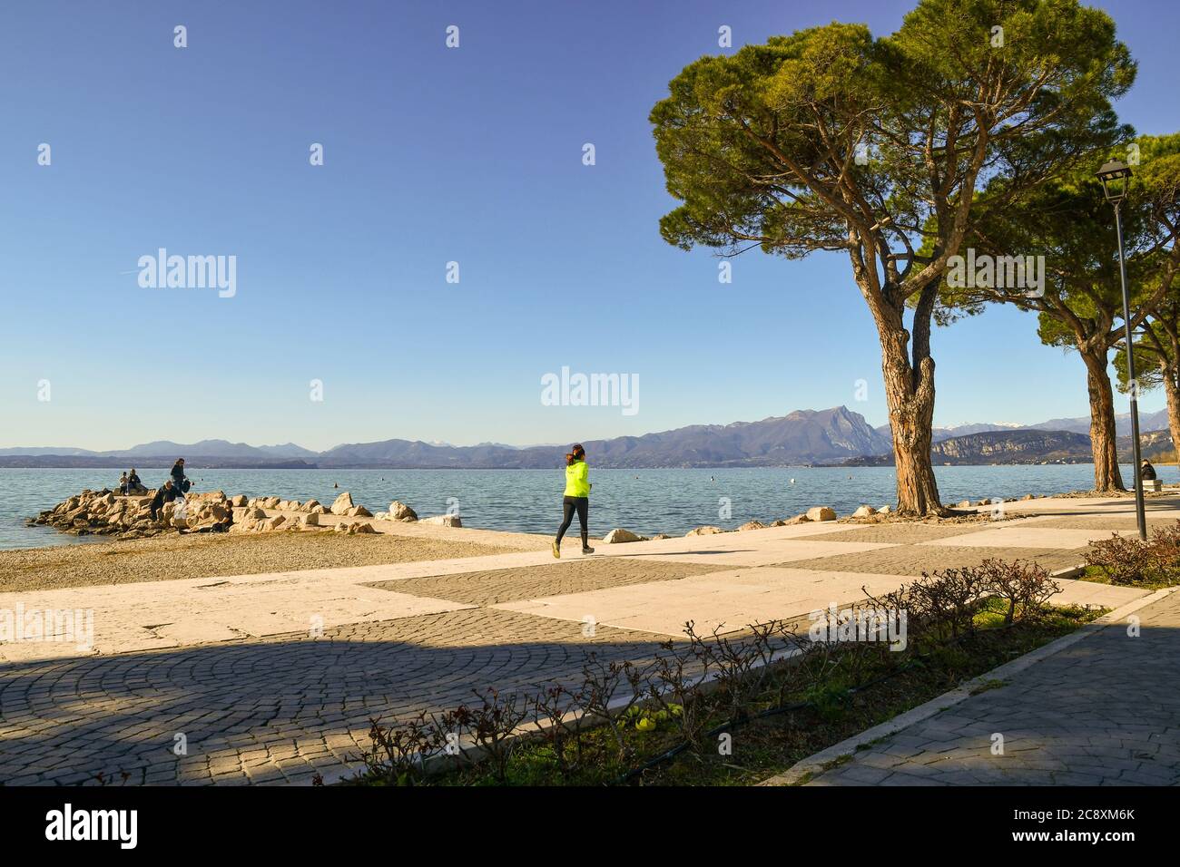 Vista panoramica del Lago di Garda con una donna che corre sul lungolago e persone sulle rocce in una soleggiata giornata invernale, Lazise, Verona, Veneto, Italia Foto Stock