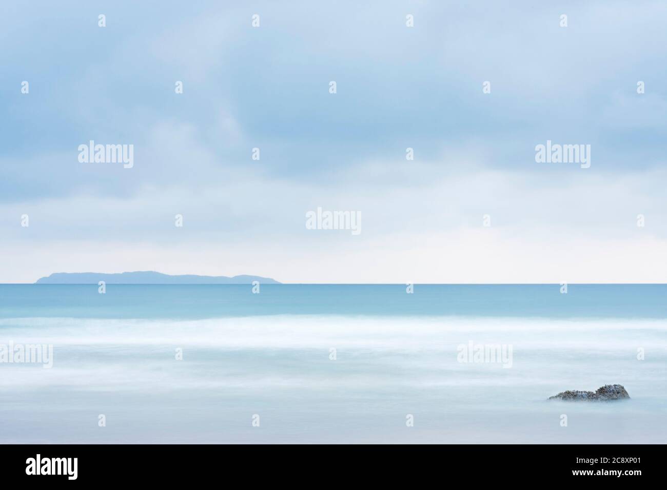 Scena tranquilla di una spiaggia al mare con un'isola sullo sfondo e una roccia in primo piano - color pastello a causa di un lungo periodo di esposizione Foto Stock