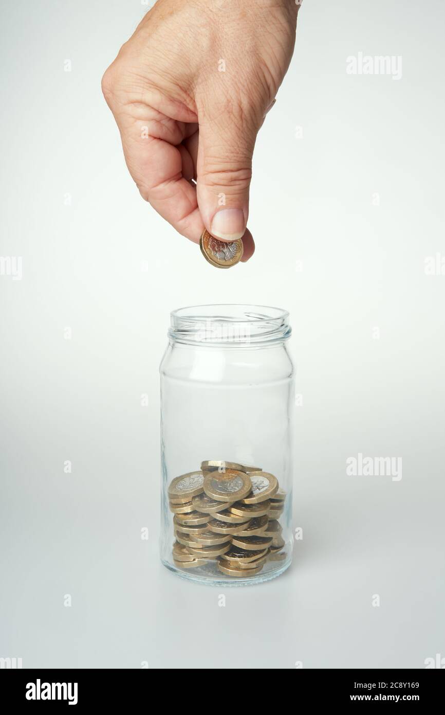 Fotografia della mano di un uomo che tiene una moneta da £1 pronta a cadere in un vaso di vetro pieno di monete. Foto Stock