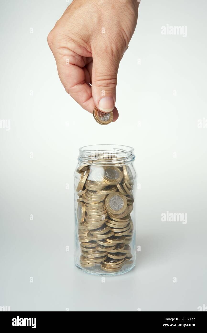 Fotografia della mano di un uomo che tiene una moneta da £1 pronta a cadere in un vaso di vetro pieno di monete. Foto Stock