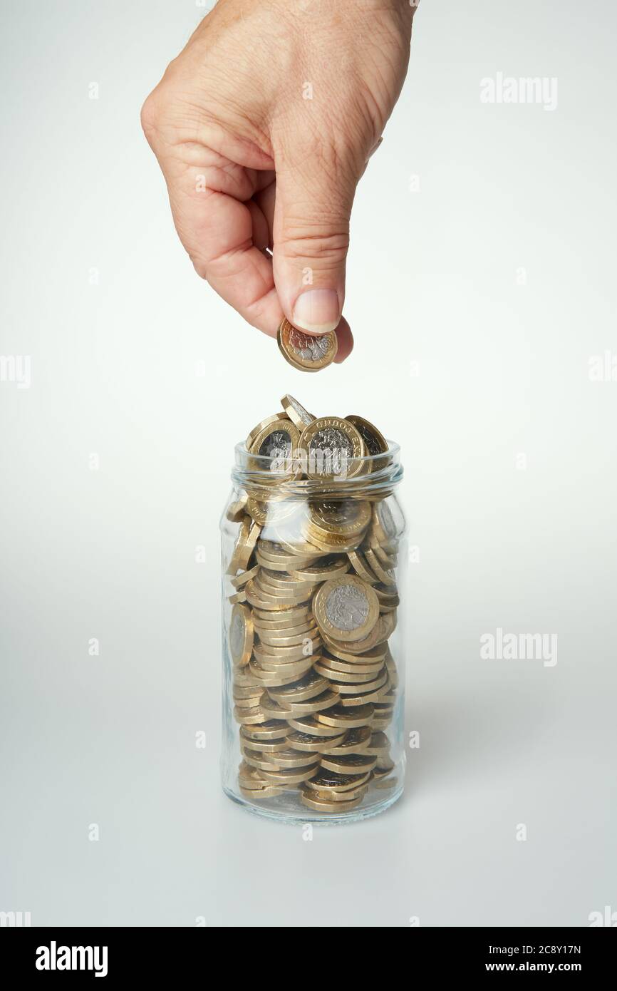 Fotografia della mano di un uomo che tiene una moneta da £1 pronta a cadere in un vaso di vetro pieno di monete. Foto Stock