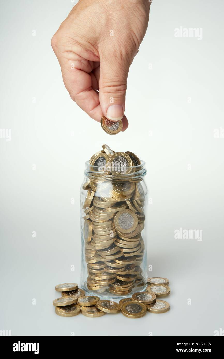 Fotografia della mano di un uomo che tiene una moneta da £1 pronta a cadere in un vaso di vetro pieno di monete. Foto Stock