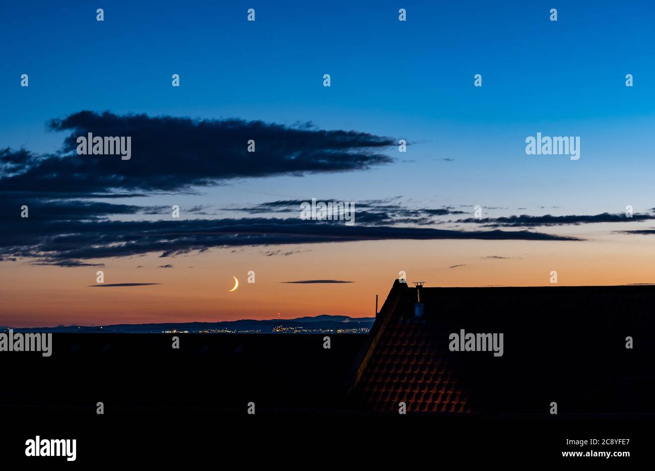 Cielo notturno estivo con una luna crescente in un tramonto arancione durante l'ora blu, vista su una silhouette di tetto a tegole, Scozia, Regno Unito Foto Stock