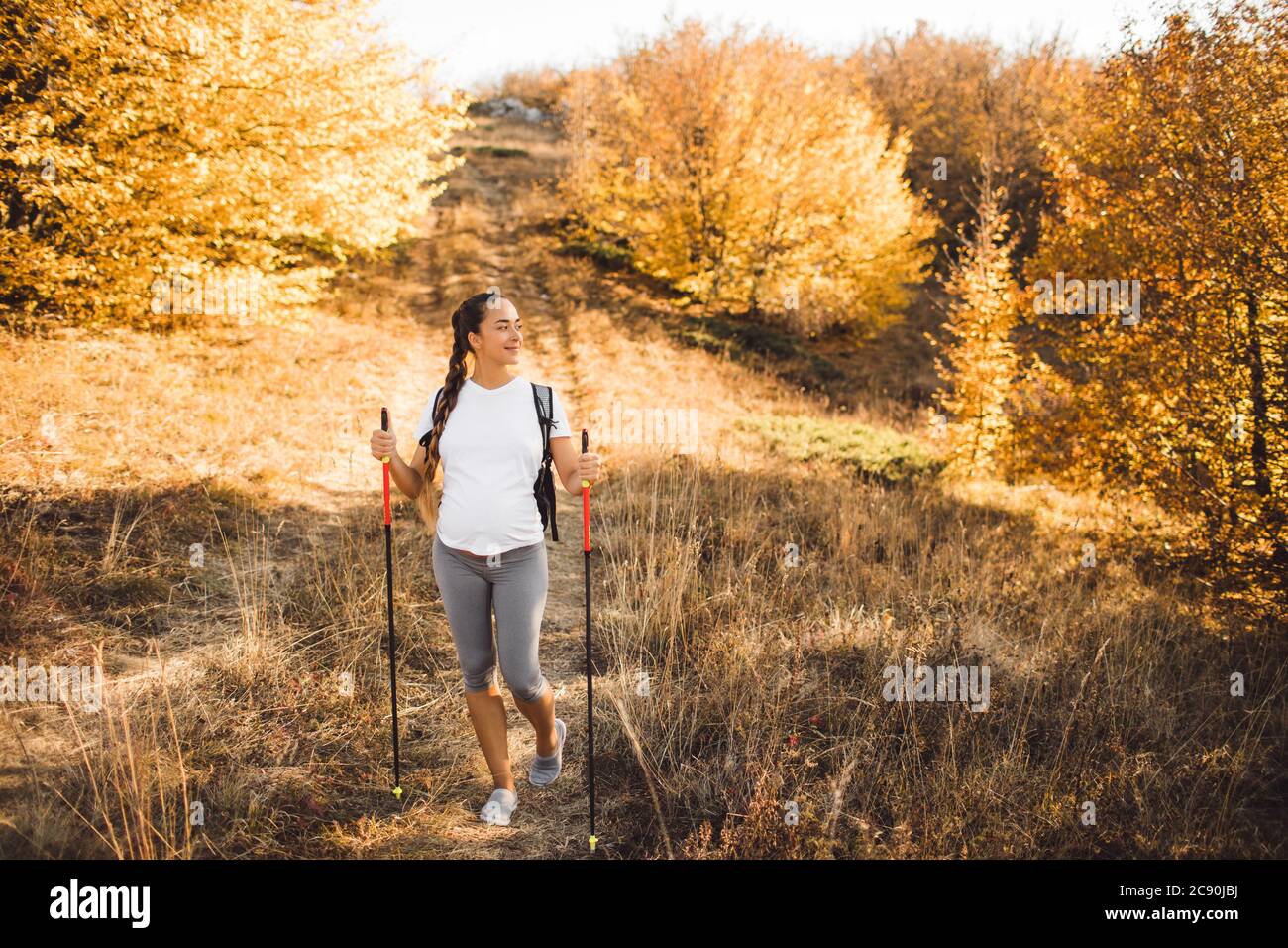 Donna incinta nordica Walking nella foresta autunnale con zaino e bastoni da trekking. Stile di vita sano e attivo nel tempo di maternità. Esercizi di gravidanza per il benessere. Foto Stock