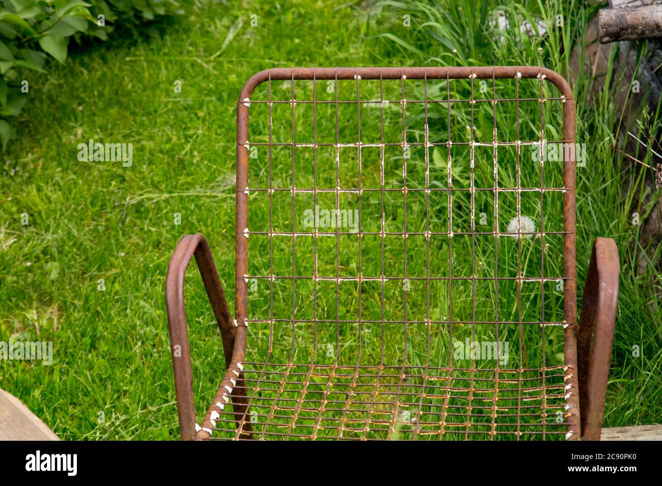 Tra qualche legno scartato, un telaio di sedia in metallo arrugginito è visto da vicino, con un sedile a rete e la schiena, di fronte all'erba verde in primavera. Foto Stock