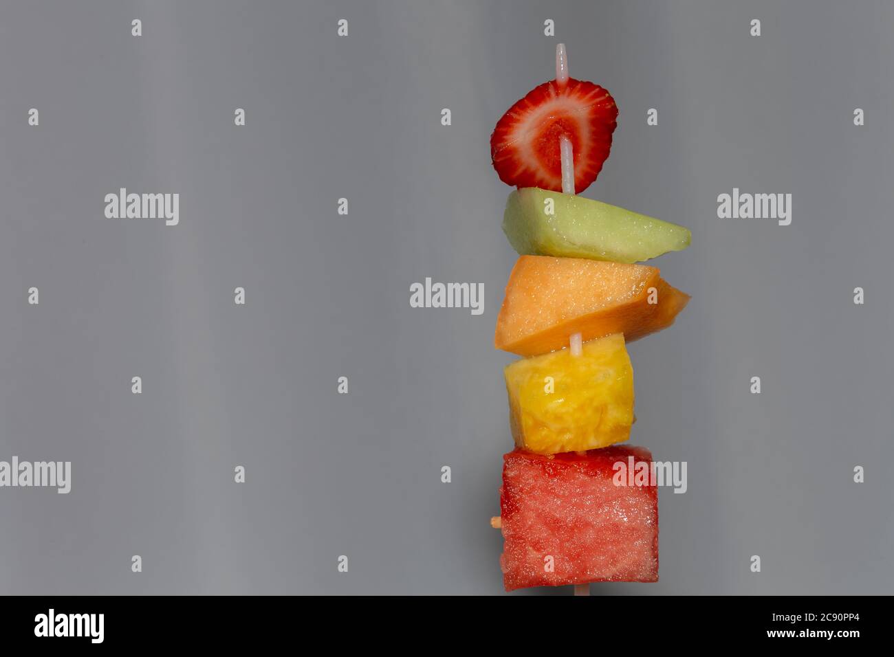 Uno spiedino di plastica ha tagliato pezzi di frutta succosa: Fragola, melone di miele, cantalupe, ananas e anguria, ed è tenuto in su prima di un backgr bianco Foto Stock