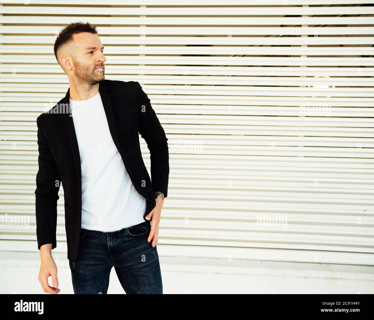 giovane uomo in giacca scura e t-shirt bianca, con barba corta che guarda  lateralmente su uno sfondo a righe Foto stock - Alamy