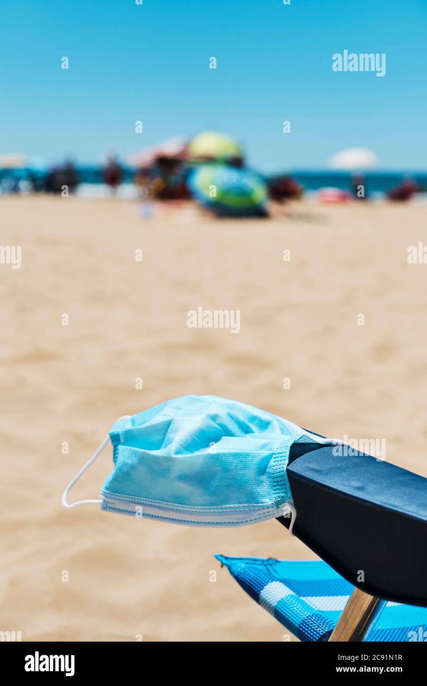 primo piano di una maschera chirurgica sul bracciolo di una sedia blu ponte sulla spiaggia, con l'oceano sullo sfondo Foto Stock