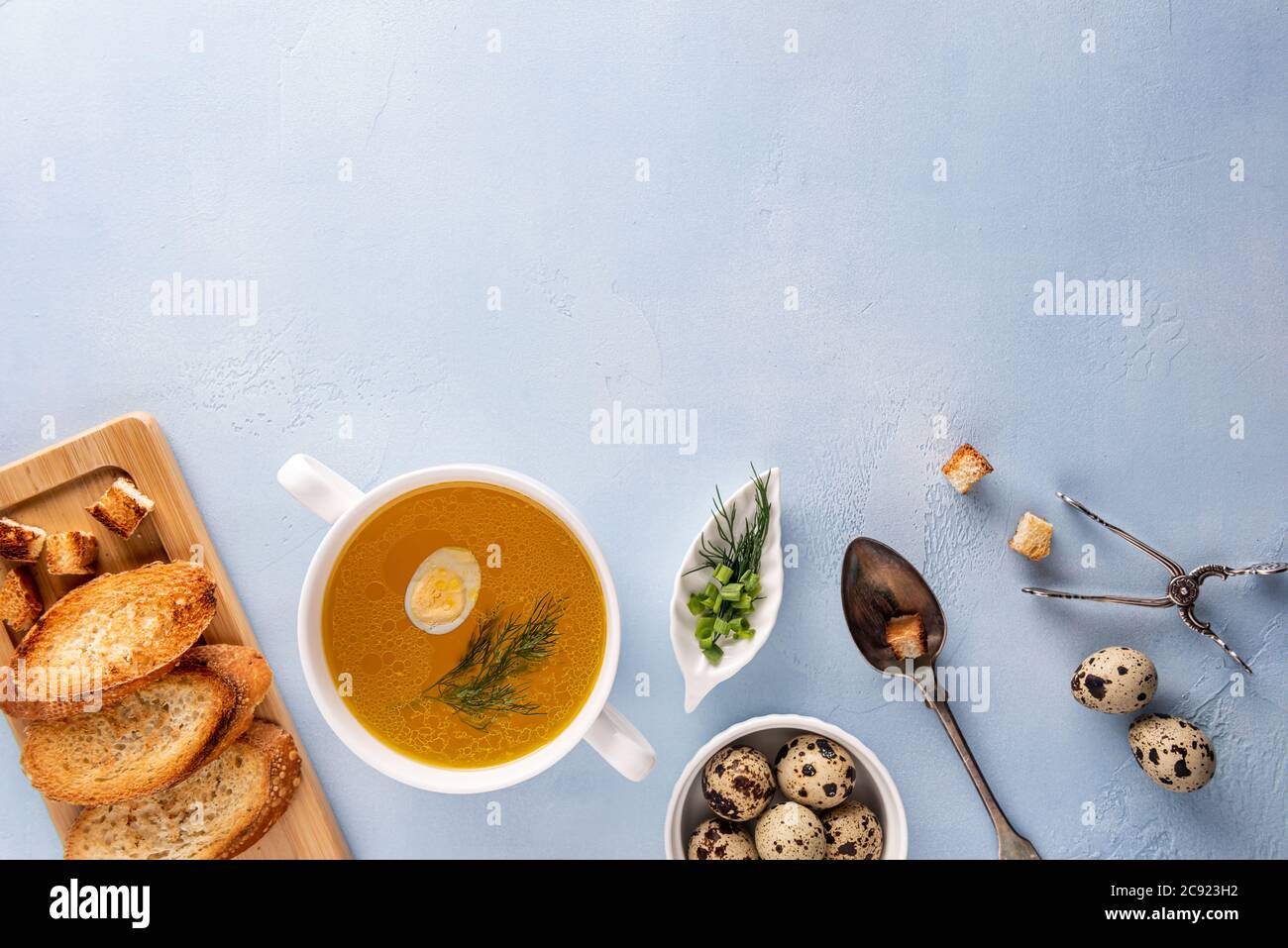 Bouillon di pollo con aneto e uova di quaglia in ciotola, toast di baguette e crostini su tagliere, cucchiaio e pinze. Piatto su sfondo blu. Grande c Foto Stock