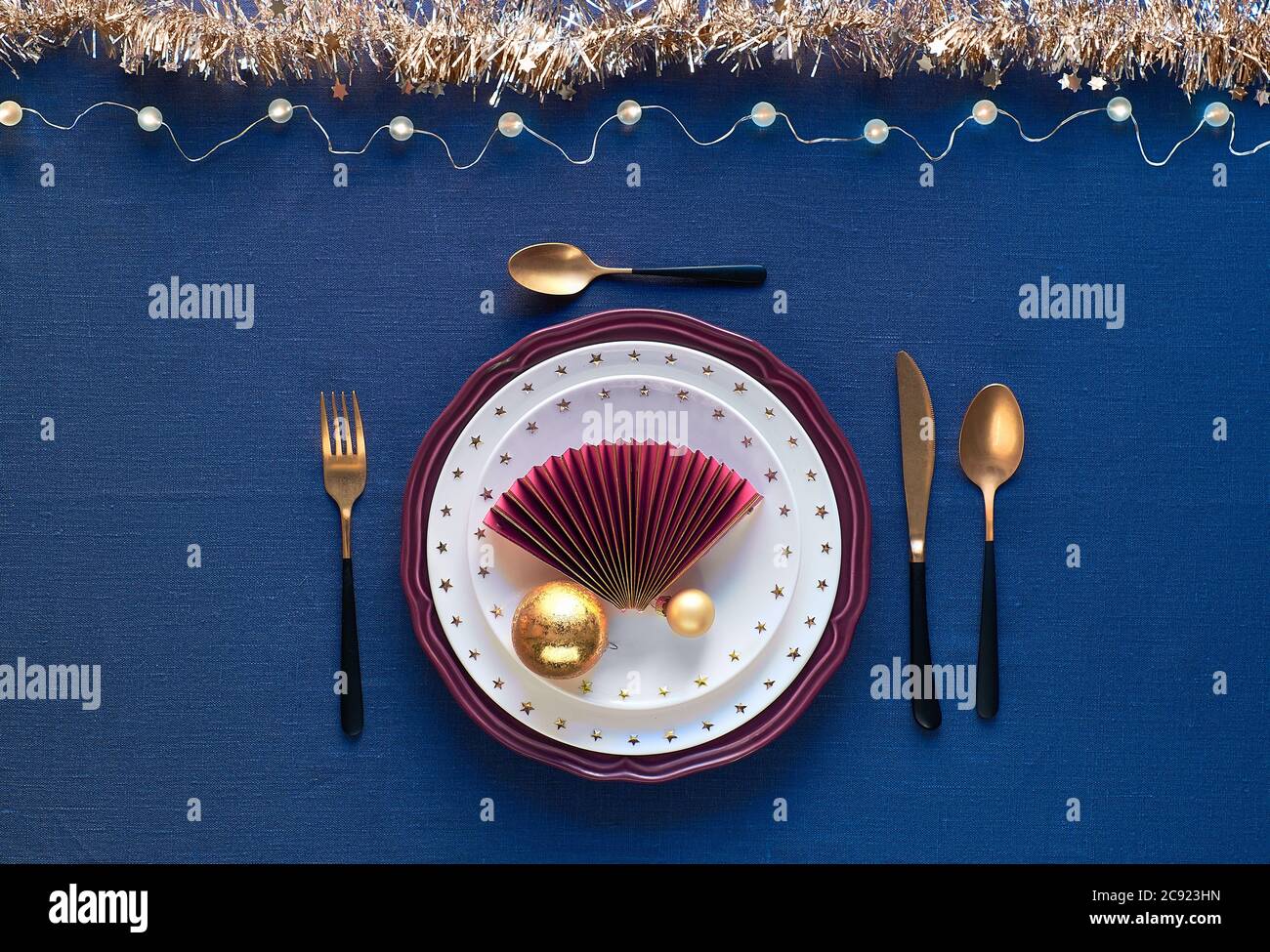Tavolo di Natale in oro, borgogna e blu scuro. Disposizione piatta, vista dall'alto su tavolo decorativo con posate dorate, piatti bianchi con s. Foto Stock