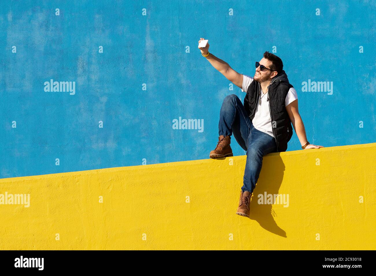 Giovane uomo con occhiali da sole seduti sulla parete gialla, prendendo selfie Foto Stock