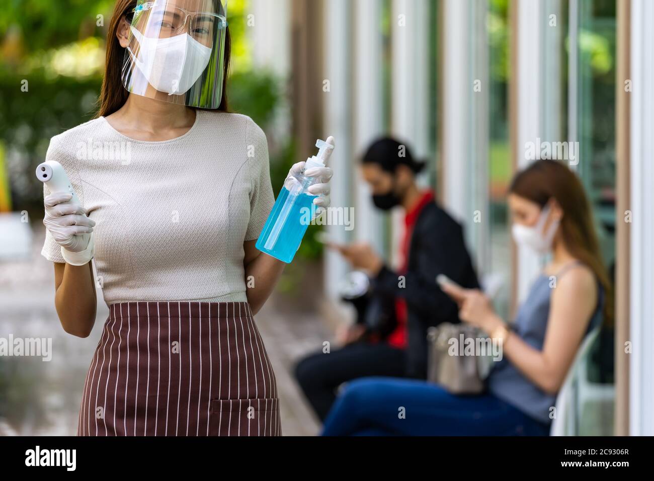 Ritratto attraente cameriera asiatica indossare maschera viso e scudo tenere termometro e igienizzatore gel di alcol con sfondo di cliente di distanza sociale Foto Stock