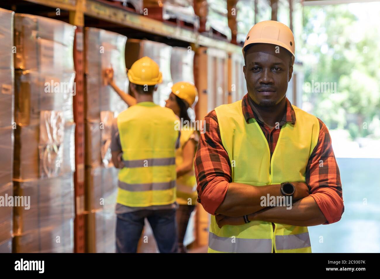 Ritratto african black warehouse supervisore in piedi e ha attraversato le braccia in un grande centro di distribuzione magazzino con lavoro di lavoro sul magazzino in dietro Foto Stock