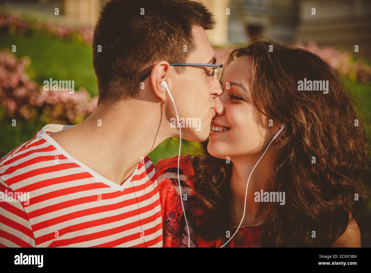 Felice coppia con gli auricolari che condividono la musica da uno smartphone in un parco all'aperto. L'uomo sta baciando la sua fidanzata Foto Stock