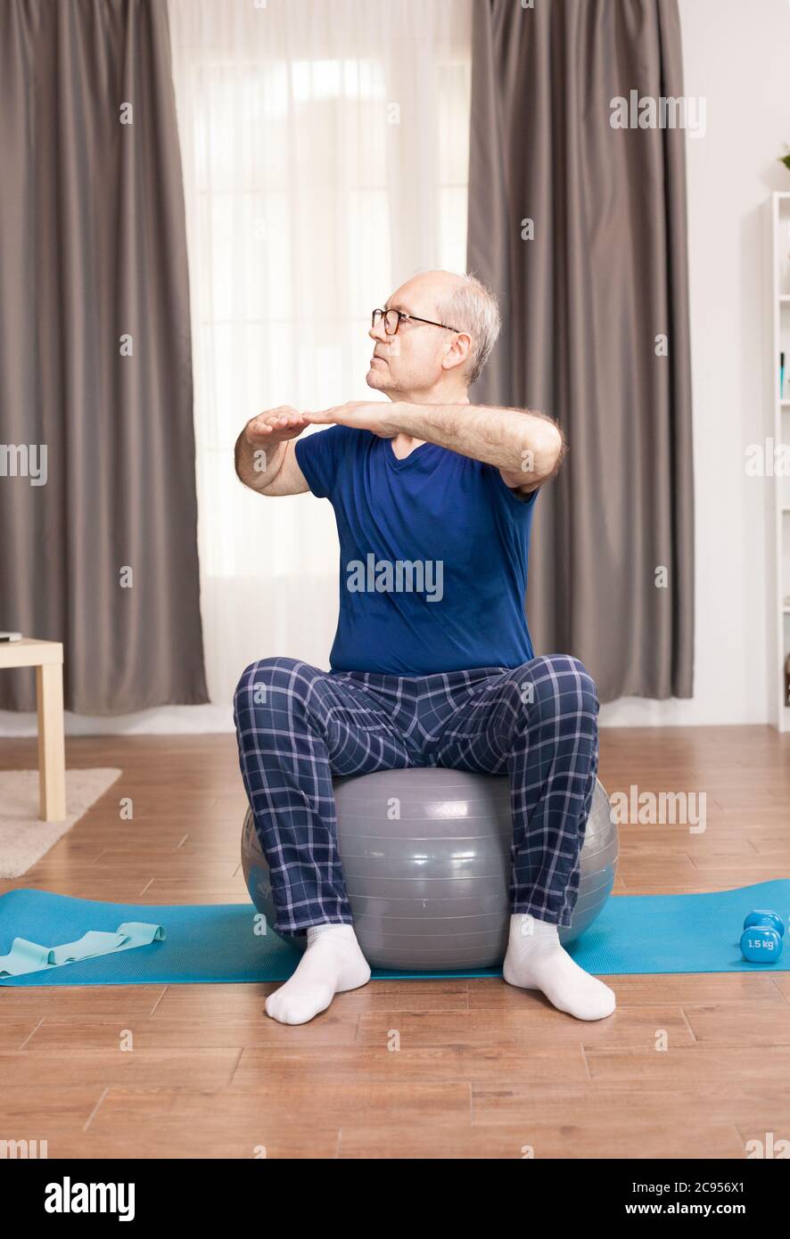 Allenamento con nonno sulla palla svizzera in soggiorno. Anziano pensionato online internet esercizio di formazione a casa attività sportiva con manubri, banda di resistenza, palla svizzera all'età di anziano pensionamento. Foto Stock