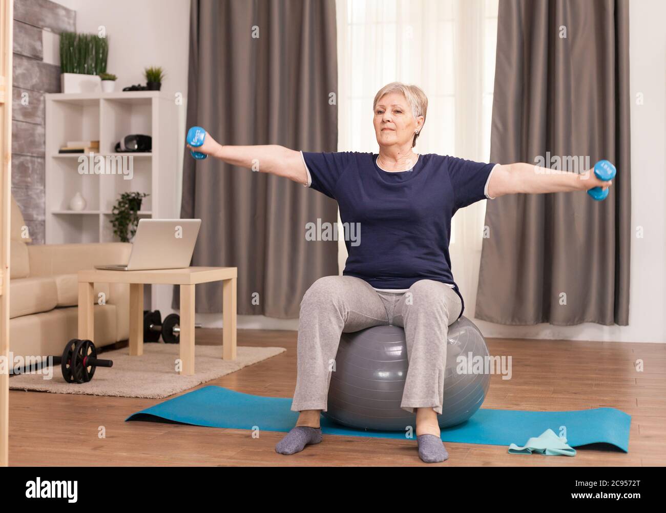 Donna anziana che fa esercizio di riscaldamento a casa. Anziano pensionato online internet esercizio di formazione a casa attività sportiva con manubri, banda di resistenza, palla svizzera all'età di anziano pensionamento Foto Stock