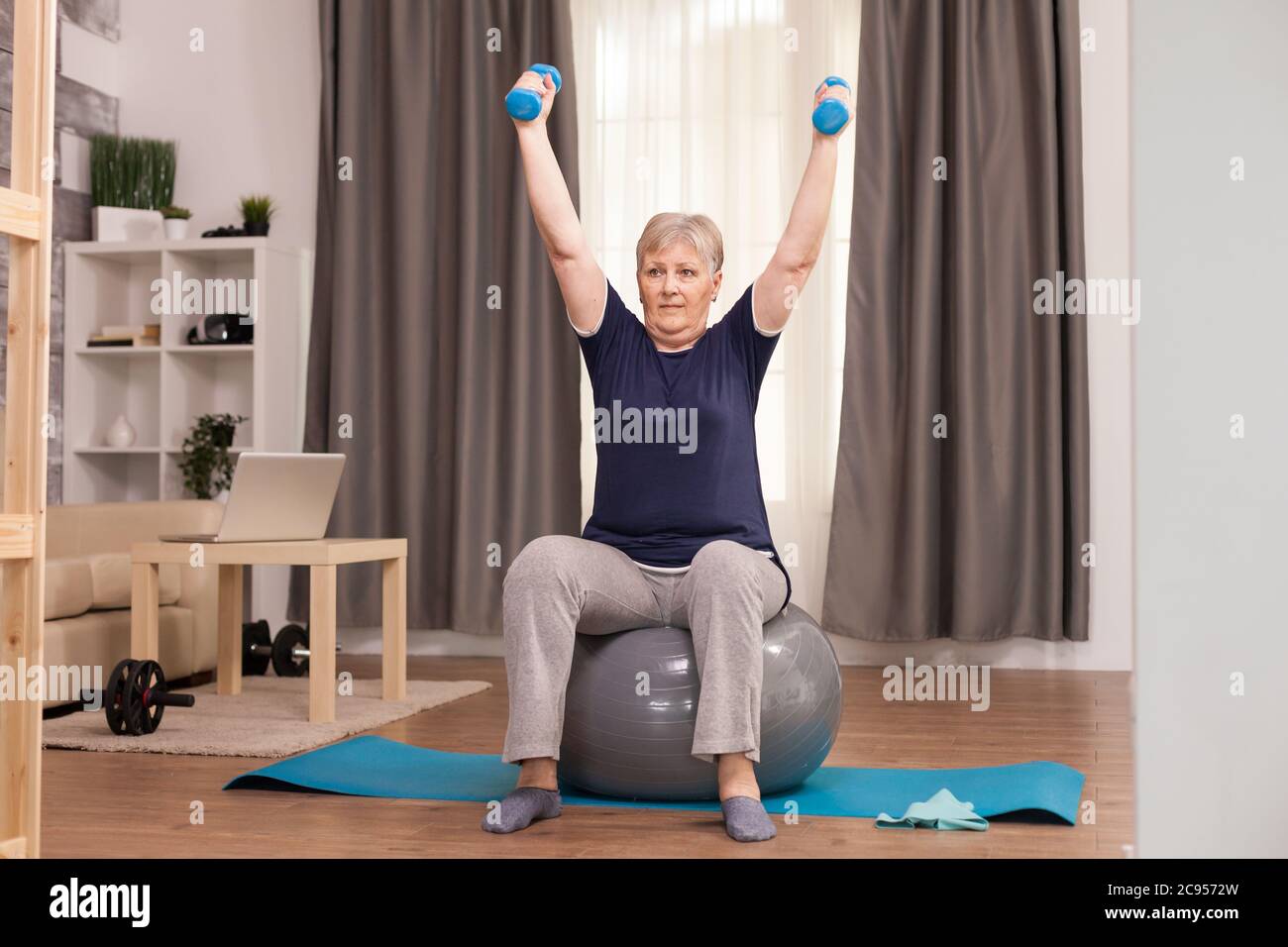 Donna anziana che fa esercizio di riscaldamento per il recupero. Anziano pensionato online internet esercizio di formazione a casa attività sportiva con manubri, banda di resistenza, palla svizzera all'età di anziano pensionamento Foto Stock