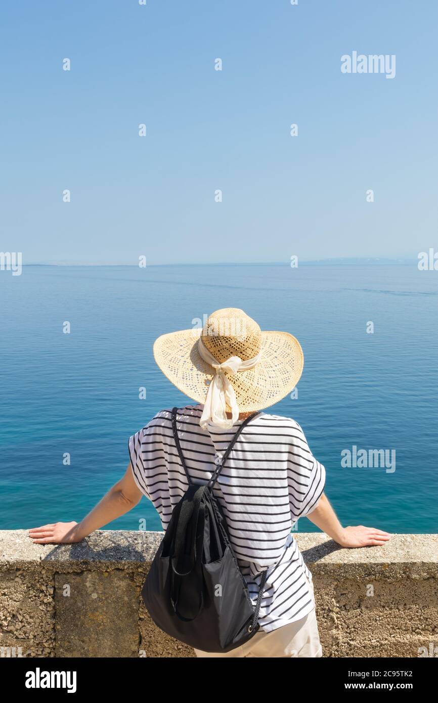 Vista posteriore di una donna viaggiatore che indossa cappello estate paglia e zaino, appoggiandosi contro un muro di pietra che guarda il mare blu grande e le isole all'orizzonte. Foto Stock