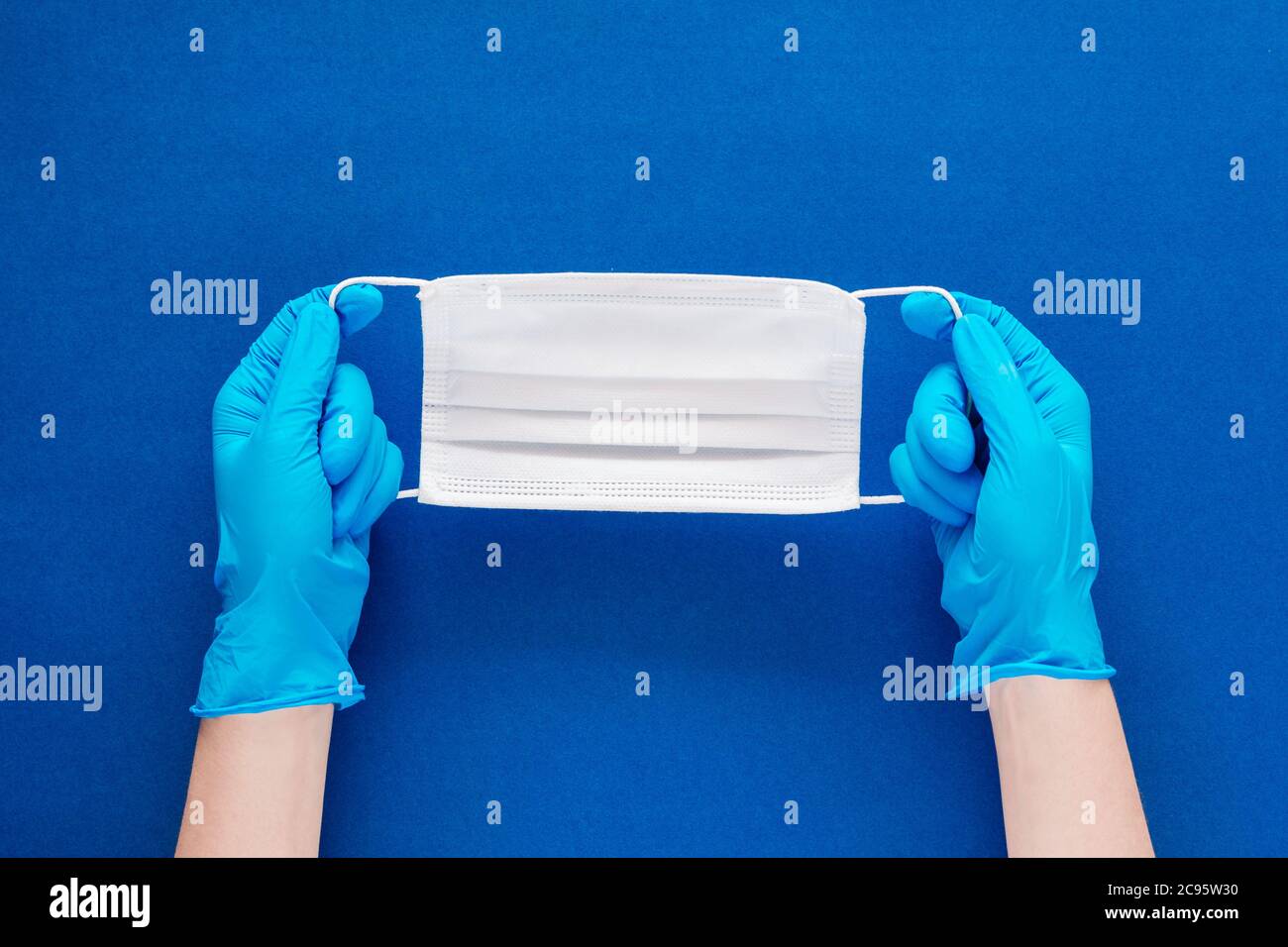 Foto in stock delle mani di una donna con guanti blu che tengono una maschera facciale su uno sfondo blu testurizzato Foto Stock