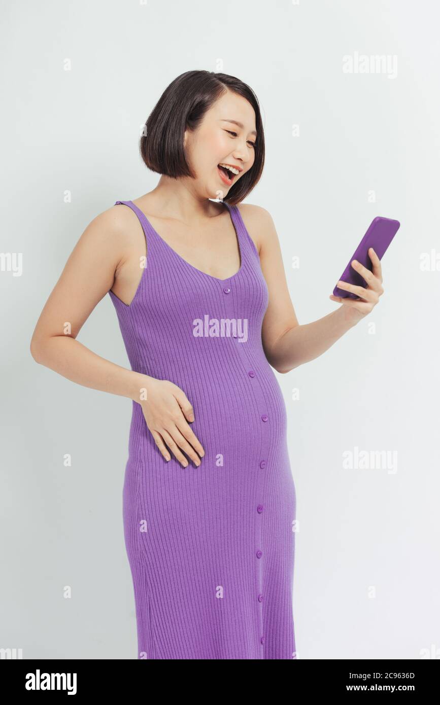 Donna incinta entusiasta che guarda i contenuti multimediali in linea su un telefono cellulare su sfondo bianco Foto Stock