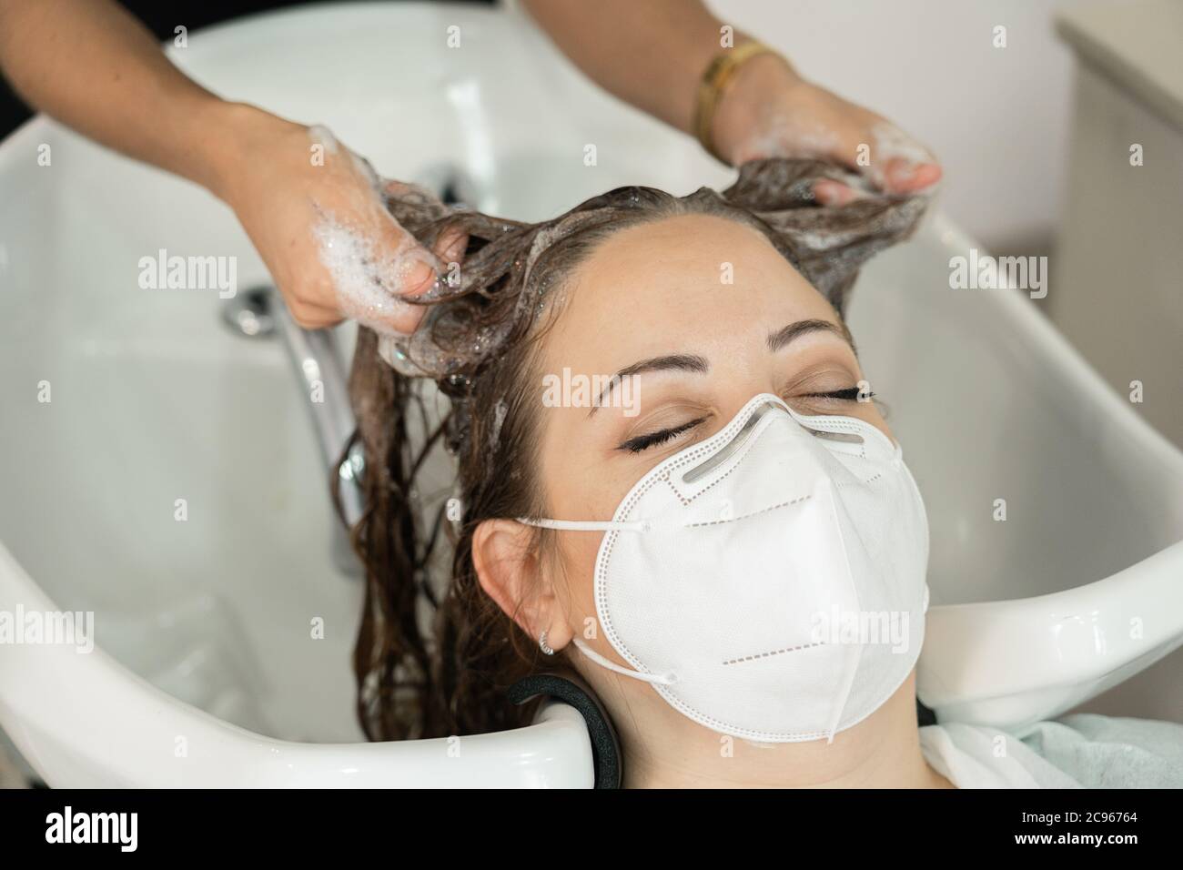 Giovane ragazza con lunghi capelli marroni, che si inzuppano lavando la testa nel parrucchiere. Distanza sociale. Uso della maschera facciale. Primo piano Foto Stock