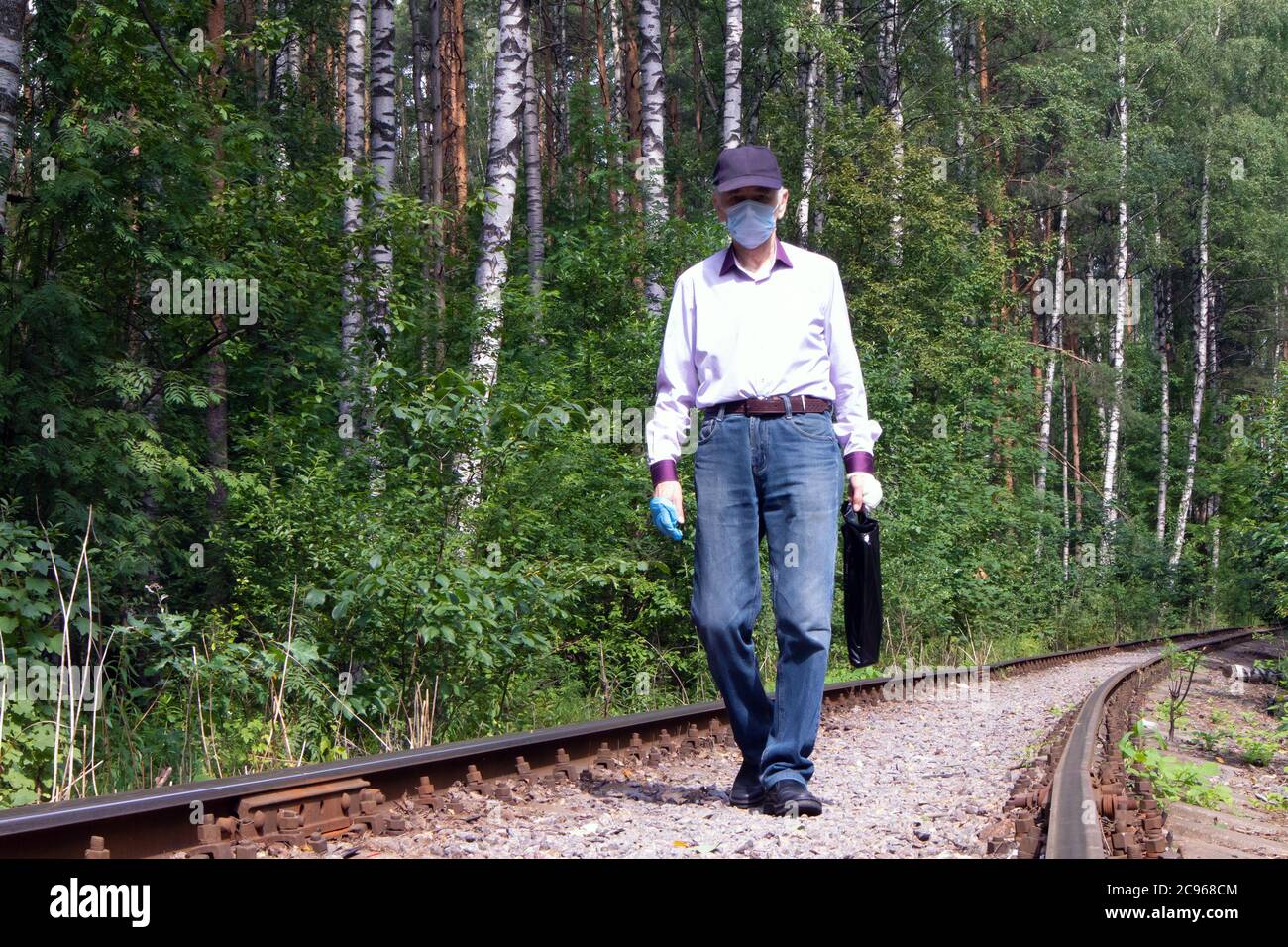 Uomo anziano in maschera medica di protezione e guanti in lattice che cammina lungo la vecchia pista ferroviaria Foto Stock