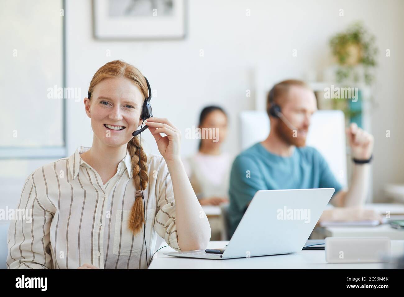Ritratto di giovane donna del servizio clienti in cuffia seduto al tavolo con il computer portatile e sorridente alla fotocamera in ufficio Foto Stock