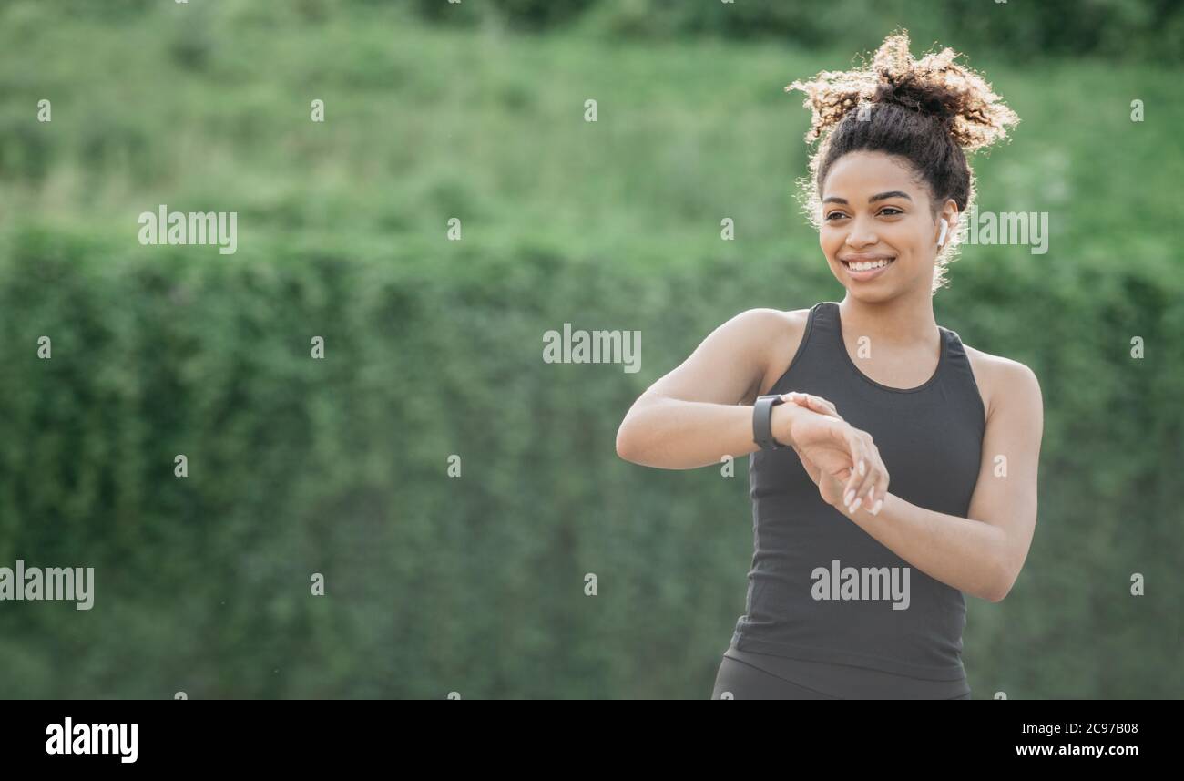 Stile di vita sano e controllo della frequenza cardiaca. Sorridente afroamericana ragazza in abbigliamento sportivo con cuffie wireless guardando fitness tracker Foto Stock