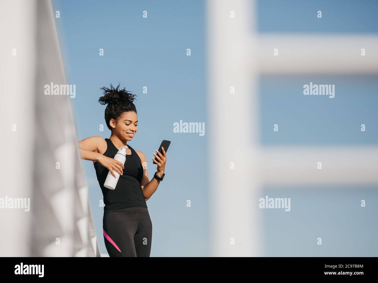 Social media e sport. Donna sorridente in abbigliamento sportivo di moda che tiene una bottiglia d'acqua e guarda lo smartphone Foto Stock