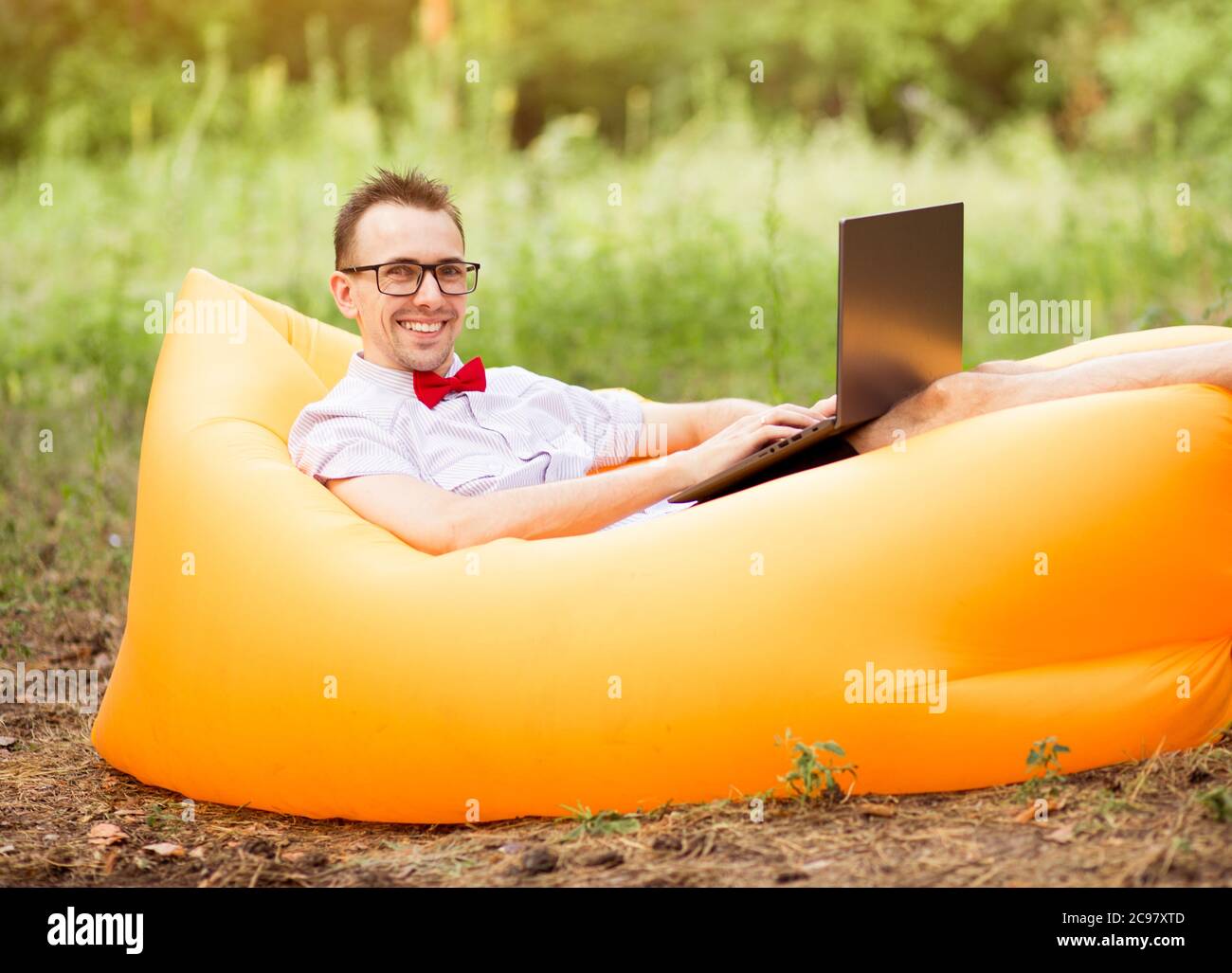 Uomo in camicia, cravatta rossa e occhiali seduti sul divano gonfiabile con computer portatile nel parco estivo. Concetto di freelancing, telelavoro Foto Stock