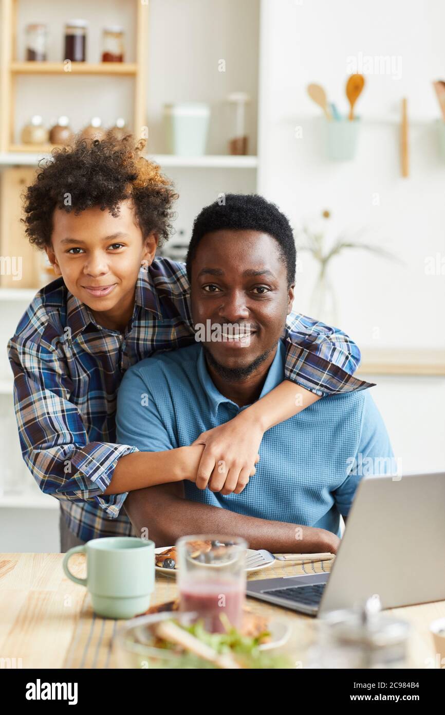 Ritratto di felice padre e figlio sorridenti alla macchina fotografica mentre si siede al tavolo con il computer portatile in cucina Foto Stock