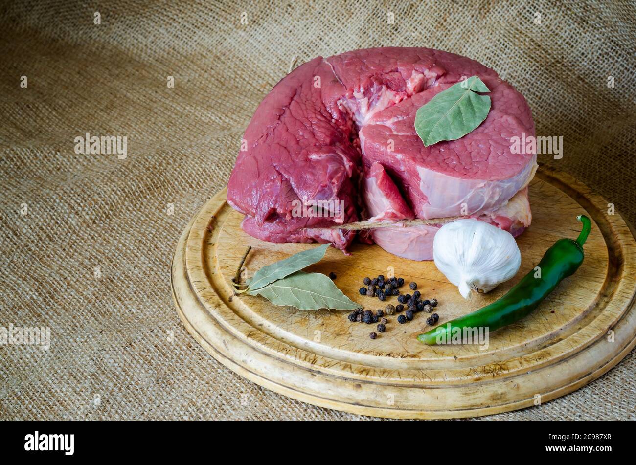 Un pezzo di manzo rotondo grande e non cotti sul burlap con aglio e spezie. Carne cruda con peperoni caldi, aglio, pepe nero e foglie di alloro su un tagliere Foto Stock