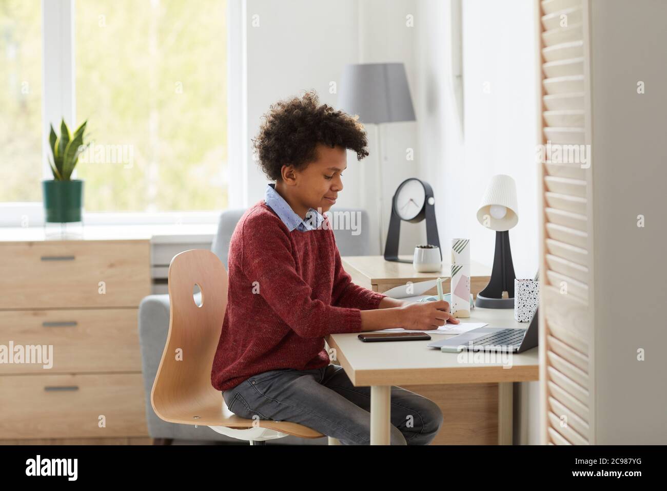 Scolaro africano seduto alla sua scrivania facendo il suo lavoro e preparandosi per le lezioni nella stanza in casa Foto Stock