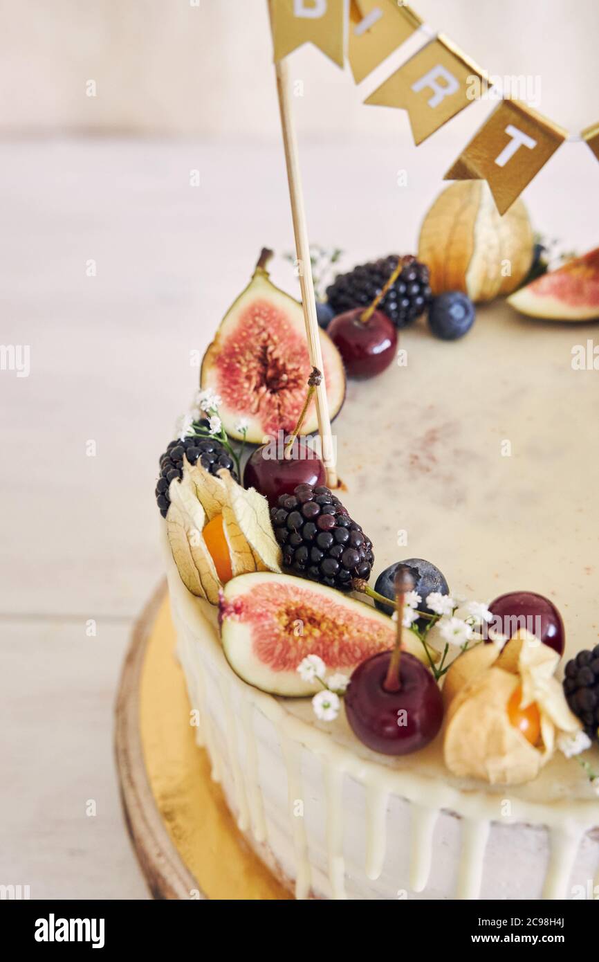 Una torta di compleanno fruttata con topper di compleanno, frutta sulla parte superiore e gocciolamento bianco su sfondo beige Foto Stock