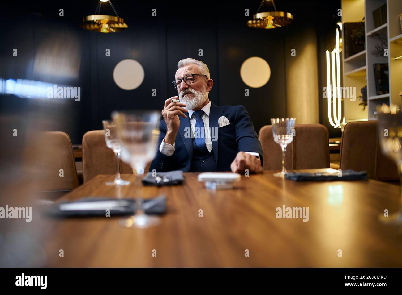 Uomo anziano dai capelli grigi che tiene e fuma e-sigaretta in un ristorante ricco. IQOS, tecnologie moderne, smetta di fumare Foto Stock