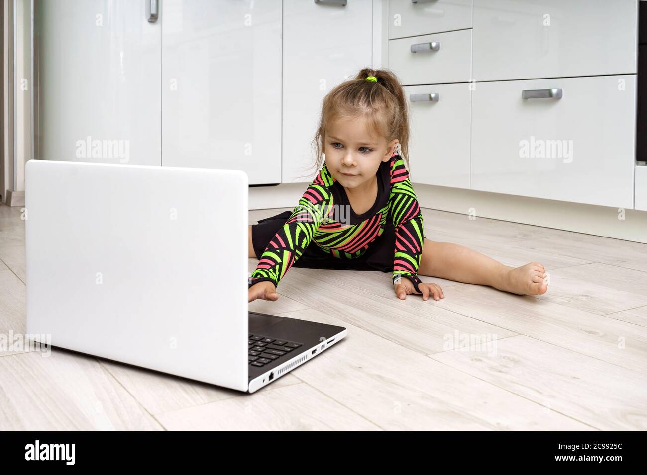 la bambina in una leotarda ginnica è impegnata in ginnastica ritmica online a casa e guarda un laptop Foto Stock