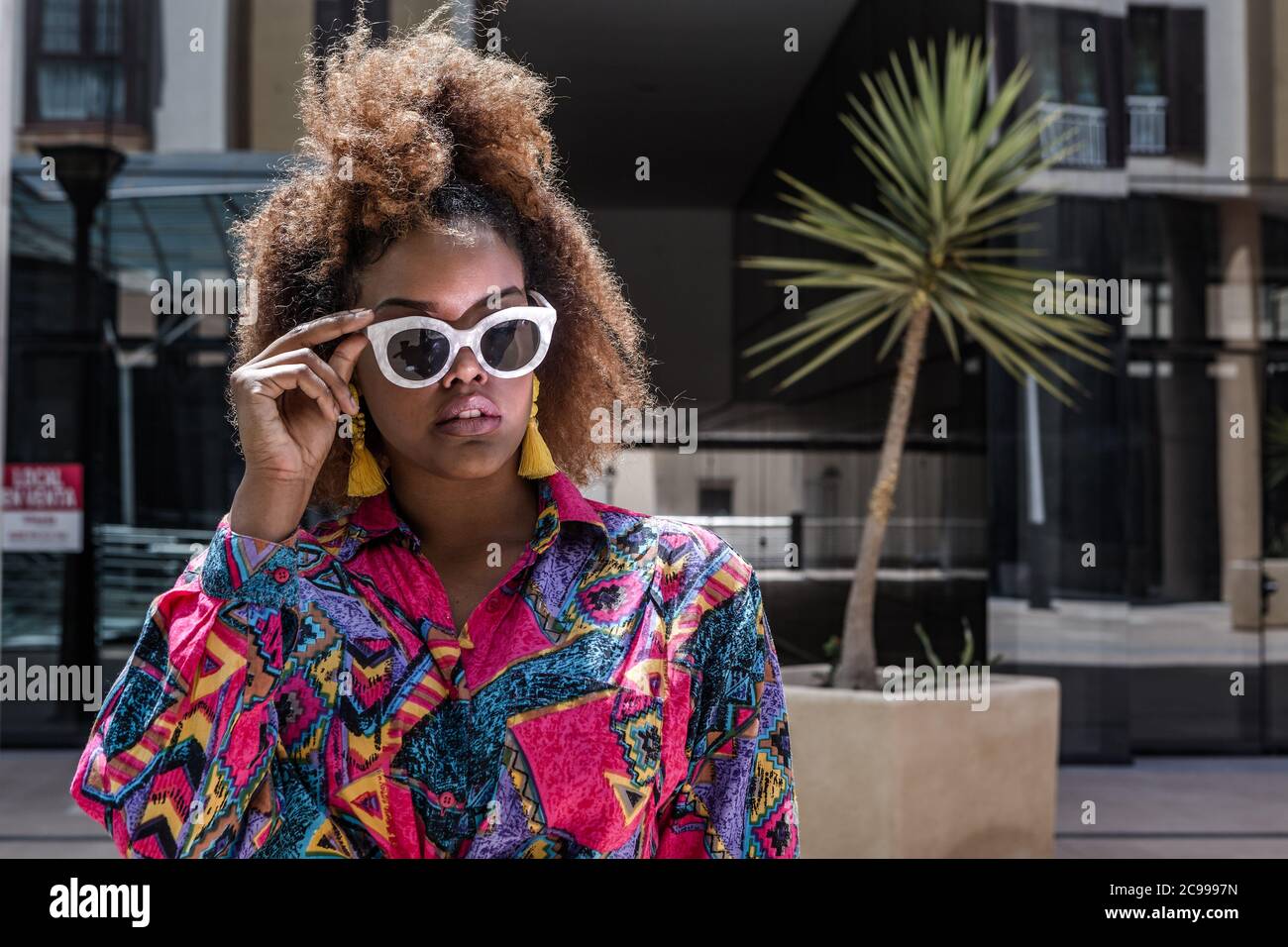 Fiduciosa millennial femminile afro-americana in abiti colorati e occhiali da sole eleganti in piedi sulla strada durante la giornata di sole Foto Stock