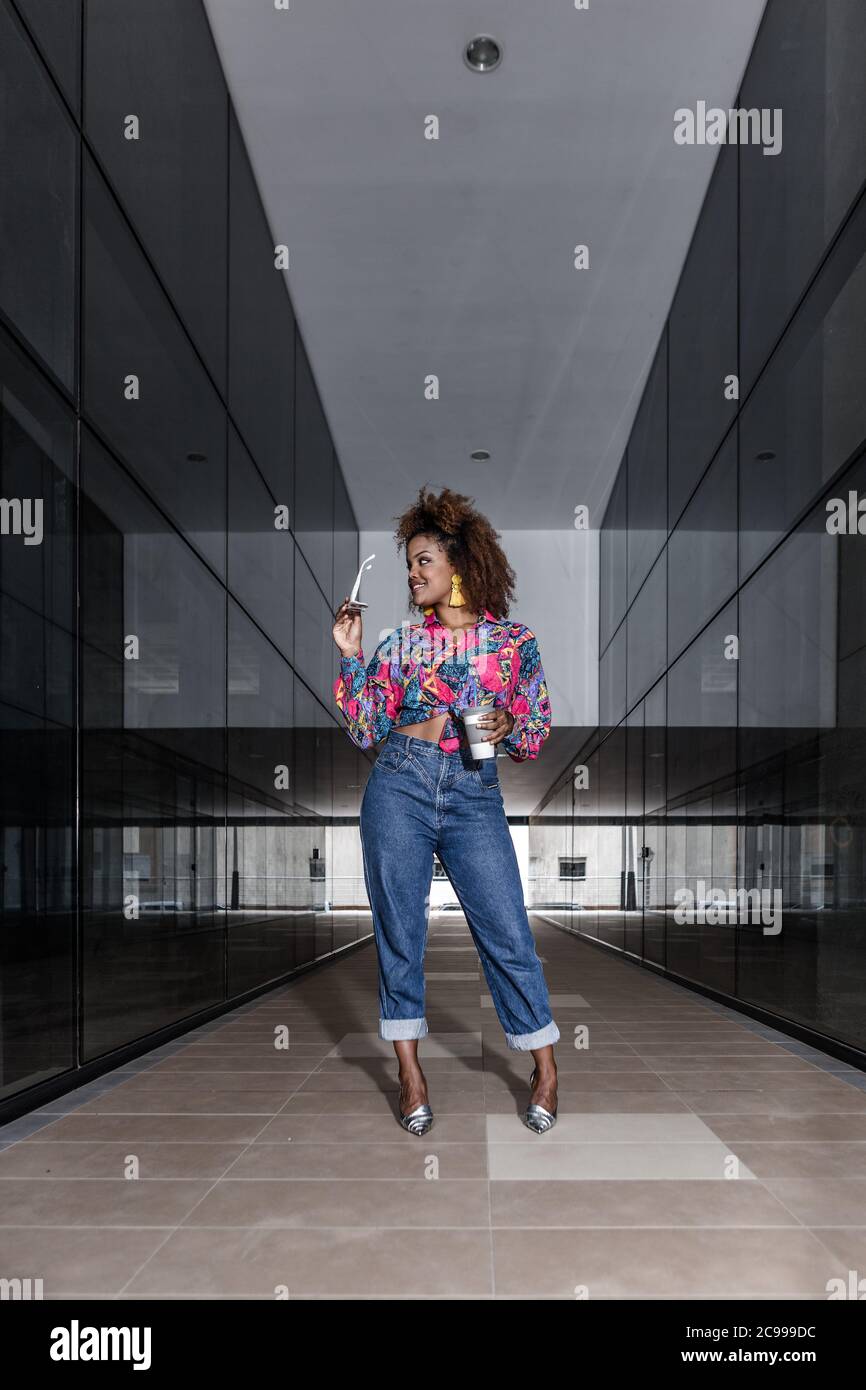 Corpo pieno di elegante donna etnica che indossa abiti alla moda in piedi con occhiali da sole e drink da asporto in città e con un'allegra vista lontano Foto Stock