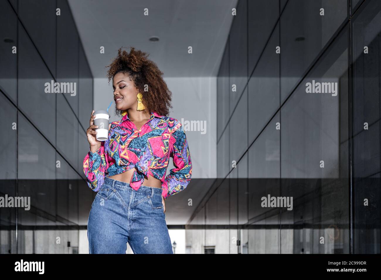 Millennial femmina nero sorridente in abbigliamento alla moda e con capelli afro in piedi con bevanda da asporto vicino a parete di vetro dell'edificio in città Foto Stock