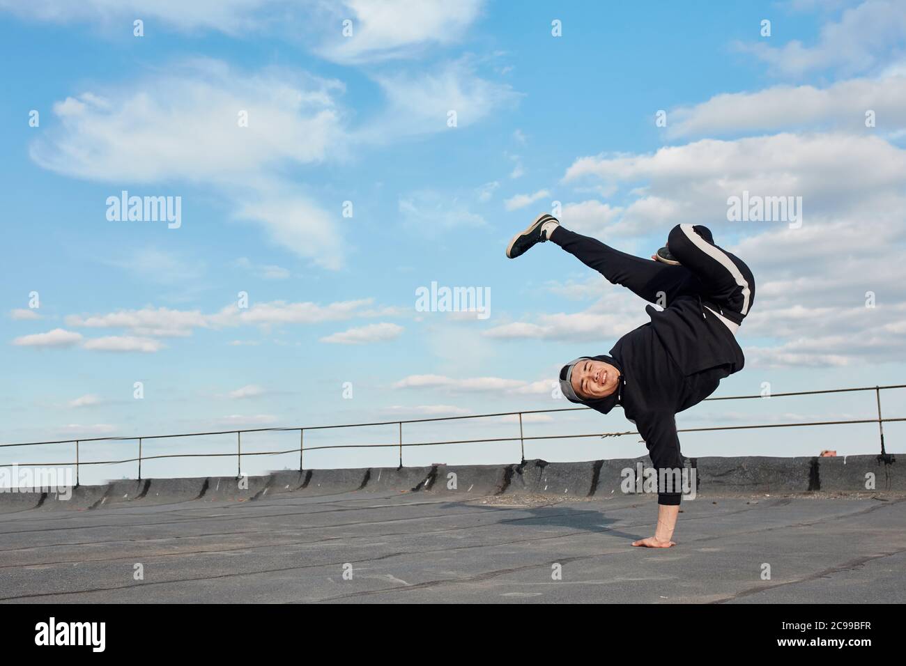 Ballerino asiatico hip-hop vestito con pantaloni sportivi neri, felpa che salta in aria, facendo un backdance acrobazie all'aperto sul tetto su sfondo blu cielo Foto Stock