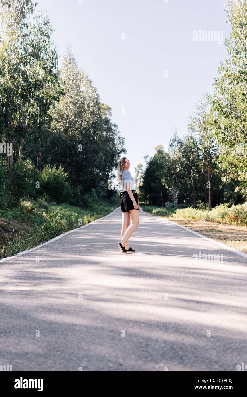 Giovane donna caucasica che cammina su una strada vuota vicino a una foresta. Foto Stock