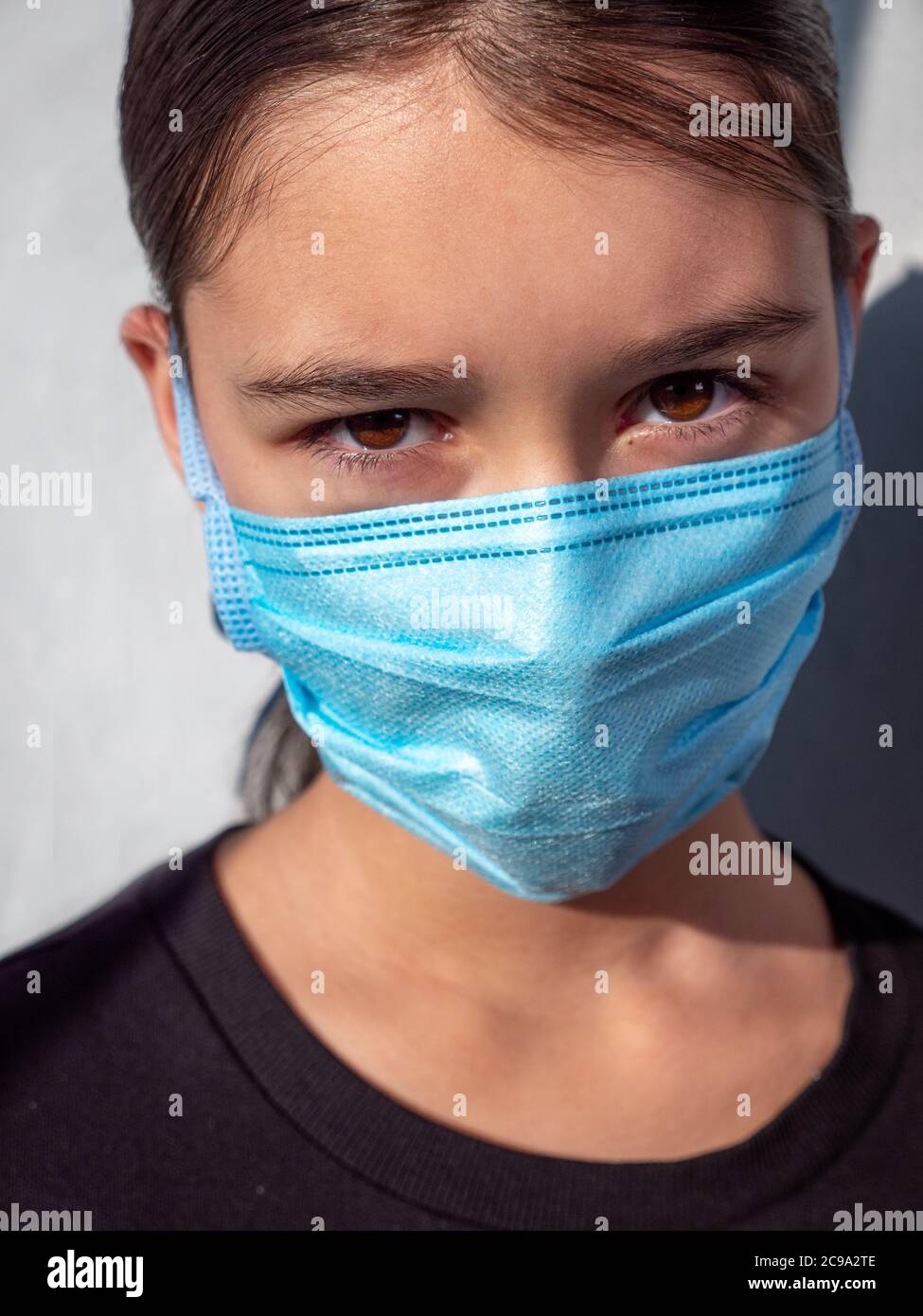 Femmina adolescente ragazza che indossa una maschera medica protettiva blu viso. Occhi noccioli e capelli castani. Protezione antivirale durante il concetto di pandemia del virus corona. Foto Stock