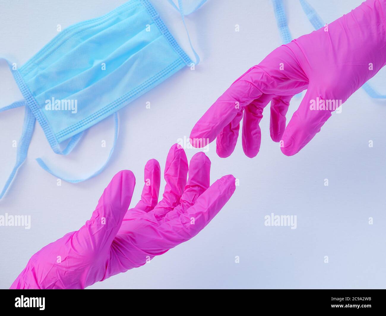 Due mani in guanti rosa in lattice che si allungano l'una verso l'altra. Maschera medica protettiva blu su sfondo bianco. Protezione antivirale durante la covid Foto Stock
