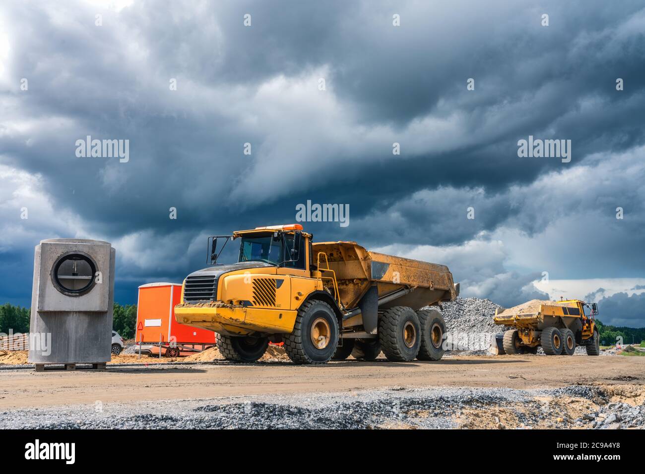 Due dumper industriali pesanti gialli parcheggiati vicino al cantiere con materiali da costruzione riforniti, nuvole blu scuro prima della pioggia. Estate soleggiata Foto Stock