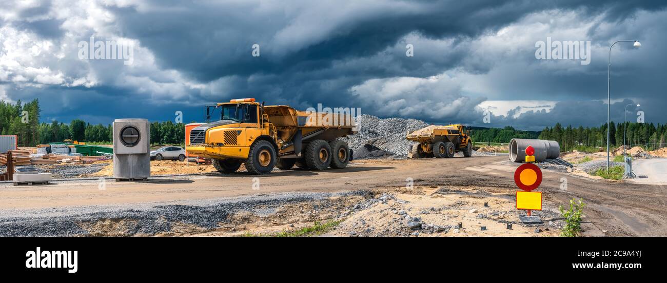 Vista panoramica su due pesanti dumper industriali gialli parcheggiati vicino al cantiere con materiali da costruzione riforniti, nuvole blu scuro prima di ra Foto Stock
