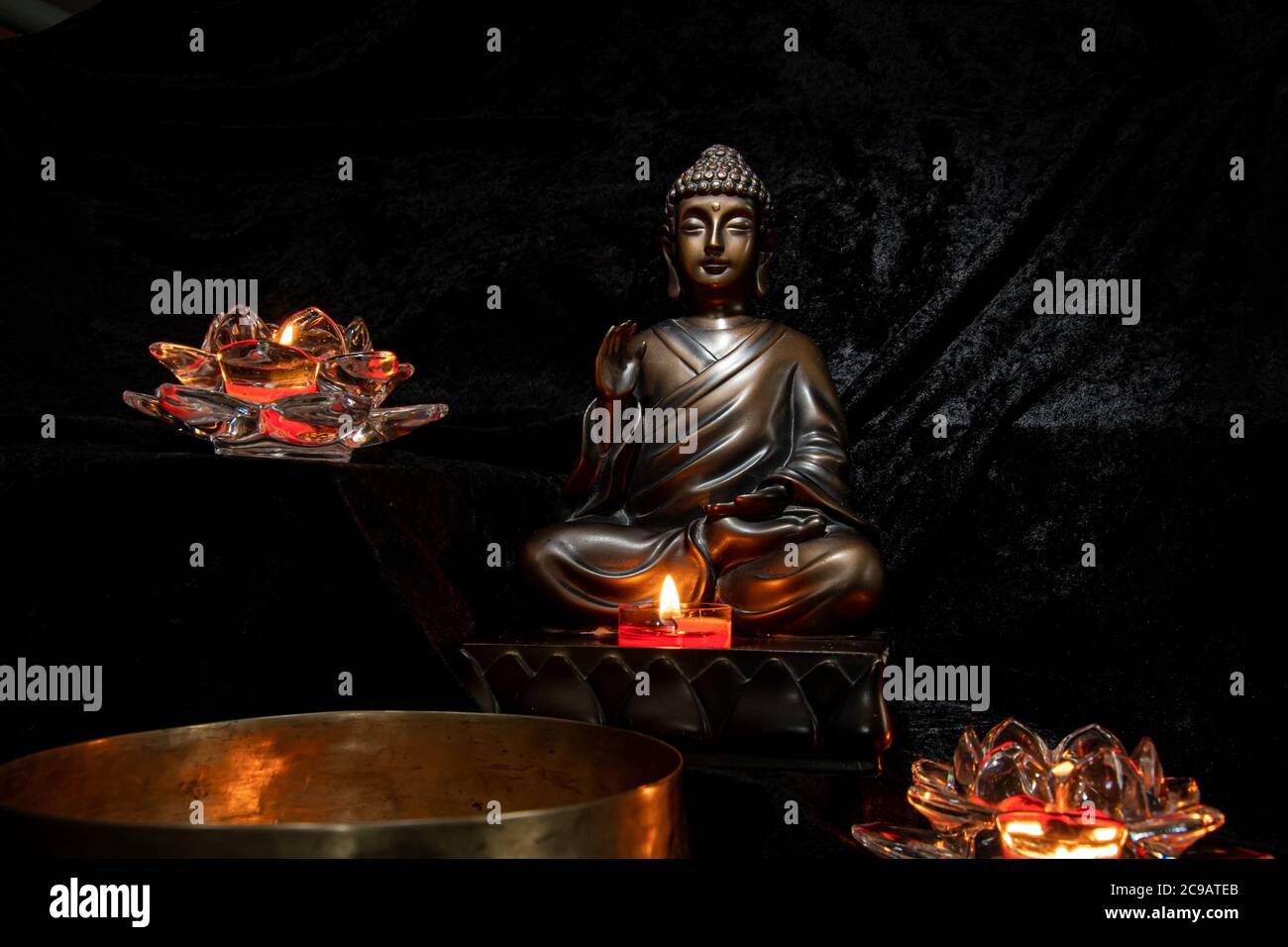 Una piccola statua del Buddha circondata da portacandele a forma di loto e con una ciotola di preghiera in ottone tibetano di fronte. Foto Stock