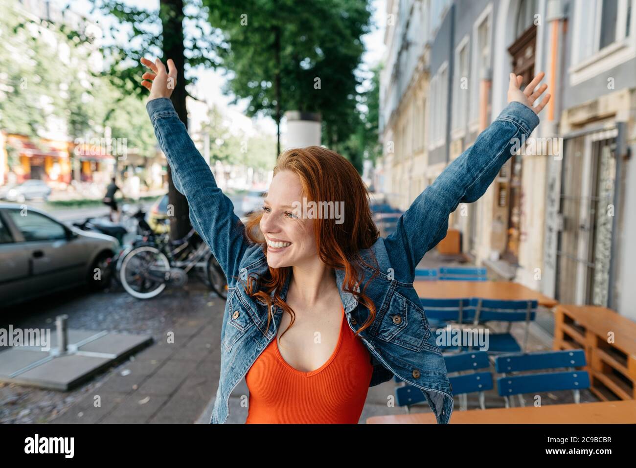 Felice casual giovane donna rossa rallegrarsi in città allungando le braccia e guardando da parte con un sorriso di piacere Foto Stock