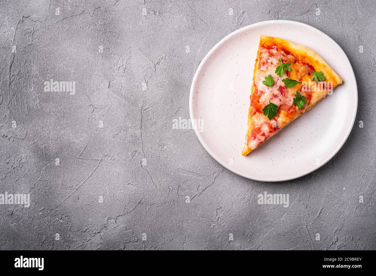 Fetta di pizza calda con mozzarella, prosciutto, pomodoro e prezzemolo sul piatto, fondo in pietra e calcestruzzo, spazio di copia per la vista dall'alto per il testo Foto Stock
